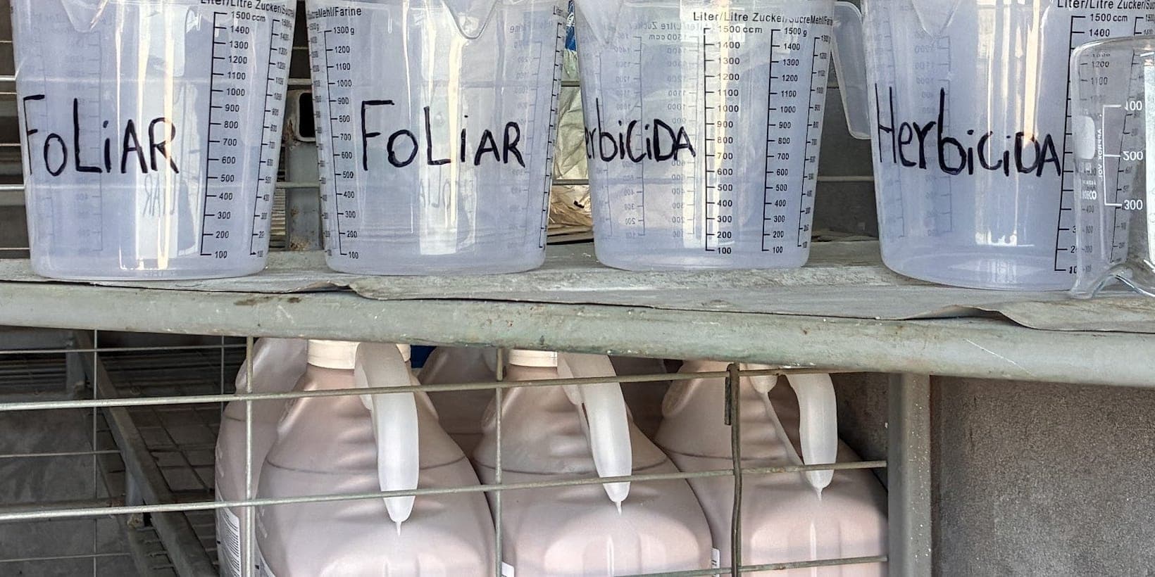 Jarros graduados rotulados por uso, foliar y herbicida, en un estante de la bodega de fitosanitarios, con bidones abajo